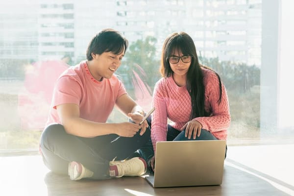 A man and a woman sitting on the floor by a window, both wearing casual pink sweaters, discussing in front of a laptop