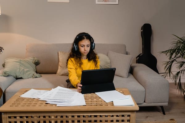 A young girl in a yellow sweater is sitting on a beige sofa, wearing headphones and using a tablet