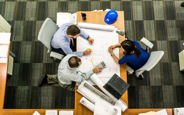 An overhead view of three people in an office setting collaborating over a large blueprint on a table