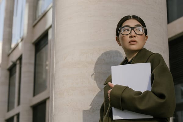 An image of a woman standing in an urban setting, wearing glasses and a green coat