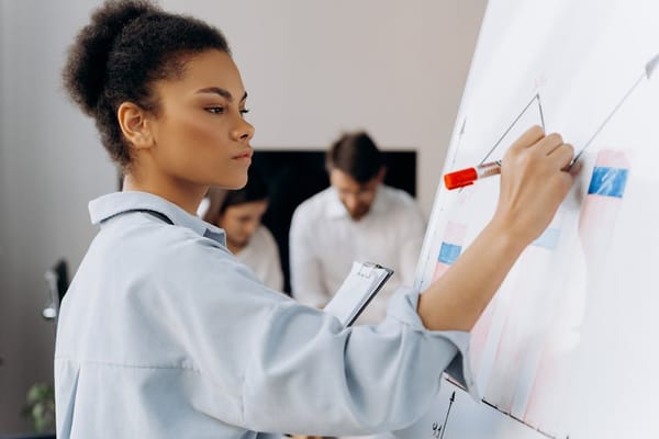 A woman in professional attire is standing near a whiteboard, drawing graphs with a red marker, indicating she's likely giving a presentation