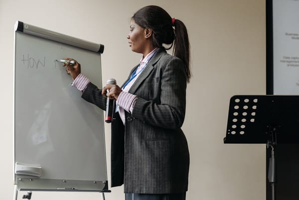 A woman in formal attire writes on a flip chart while holding a microphone, suggesting she is giving a presentation or lecture