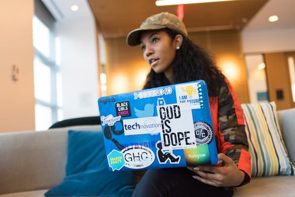 A woman wearing a camouflage jacket and cap is sitting on a couch with a colorful laptop covered in various inspirational and tech-themed stickers