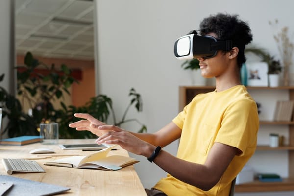 A person wearing a virtual reality headset, sitting at a desk with a keyboard, notebook, and glass of water