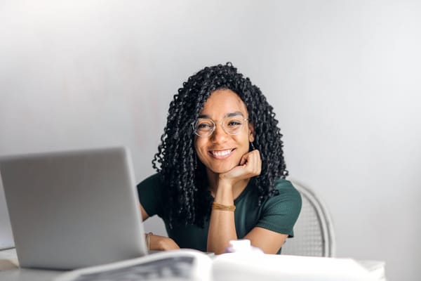 A woman with curly hair and glasses is sitting at a desk, smiling at the camera