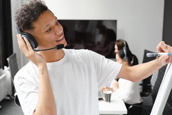 A man wearing a white T-shirt and a headset is standing in front of a whiteboard, writing or drawing with a marker