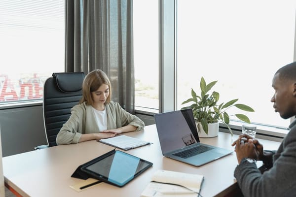 A business meeting in a modern office setting featuring a woman and man sitting across from each other at a table with laptops and documents