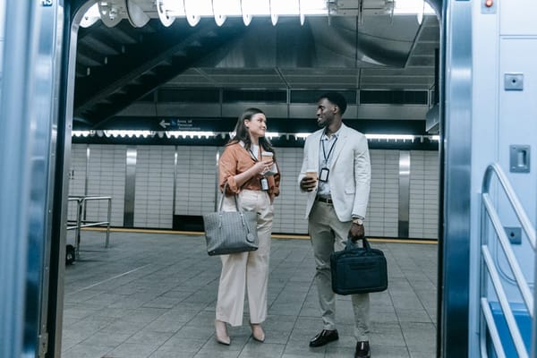 A man and a woman standing inside a subway station, holding coffee cups and looking at each other, suggesting a friendly conversation