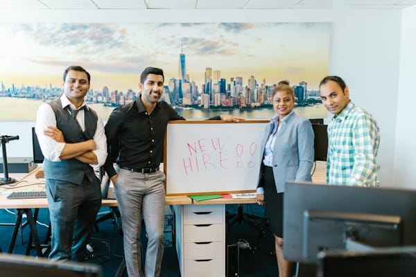 A group of four people standing in an office environment, smiling and posing around a whiteboard with the words 'New Hire' written on it